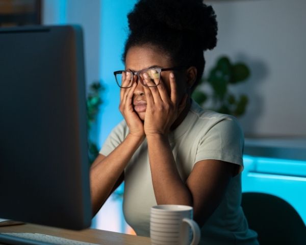 Fadiga ocular por telas: sinais, pausas e quando avaliar 1 Mulher usando computador à noite e esfregando os olhos devido ao cansaço visual, representando fadiga ocular por telas, visão embaçada e impacto do uso prolongado de telas na saúde ocular.