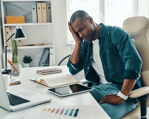 Como o esforço visual pode causar dor de cabeça 1 Homem sentado no escritório com a mão na cabeça, ilustrando dor de cabeça e problemas na visão causados por esforço visual durante o trabalho.
